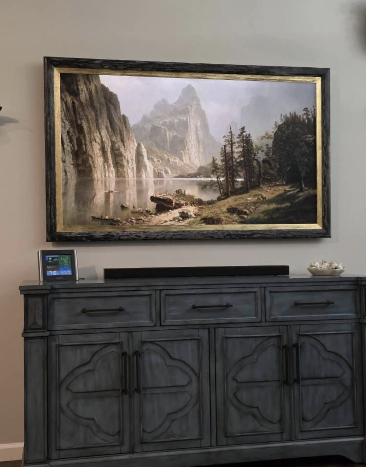 Dark TV frame with a gold inner edge displaying a mountain landscape above a carved cabinet.