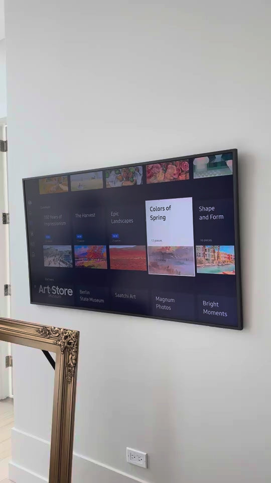 A person fitting a fully assembled gold frame around a wall-mounted TV.